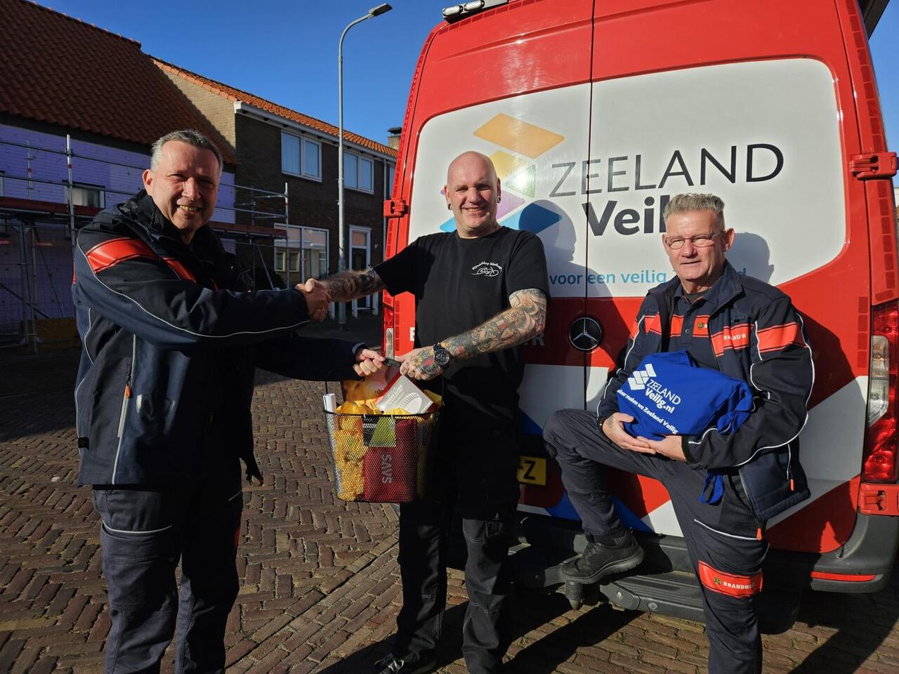 De brandweermannen Johan Koppejan (links) en Ronald van Leeuwen (rechts) overhandigen het verrassingspakket aan rijwielhandelaar Gerard Janse..